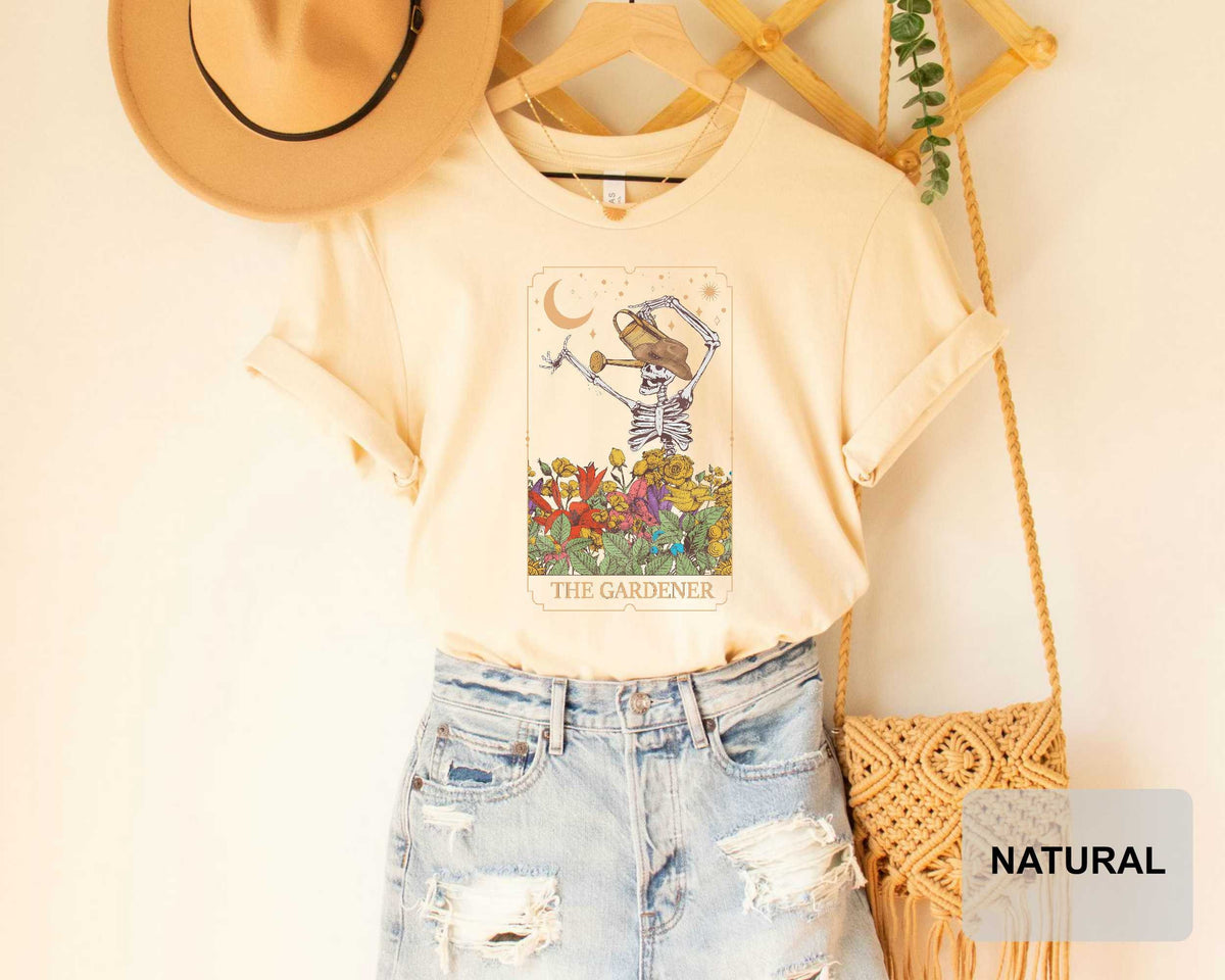 A beige t-shirt with a graphic design of a skeleton holding a flower, along with a pair of denim shorts and a straw bag, all displayed on a wooden hanger.
