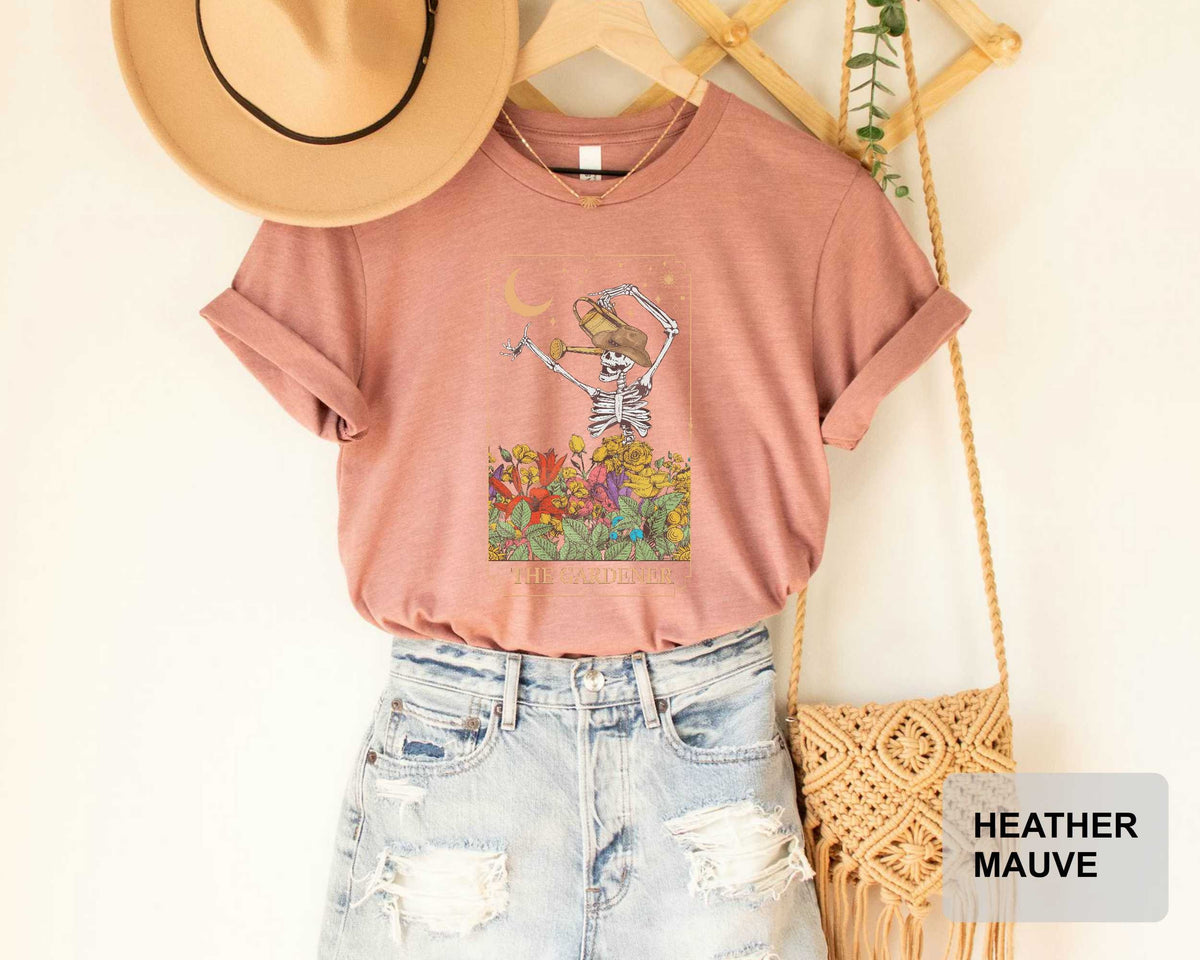 A pink t-shirt with a skeleton graphic and floral design, a beige hat, distressed denim shorts, and a woven purse are displayed on a hanger.