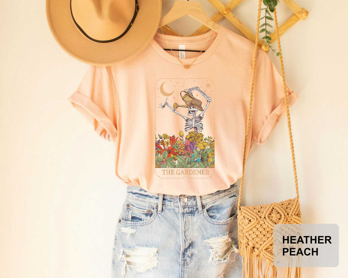 a pink t-shirt with a graphic design of a skeleton holding a flower, along with the text "THE GARDENER". The t-shirt is hanging on a wooden hanger, and there is a beige hat and a woven purse nearby.