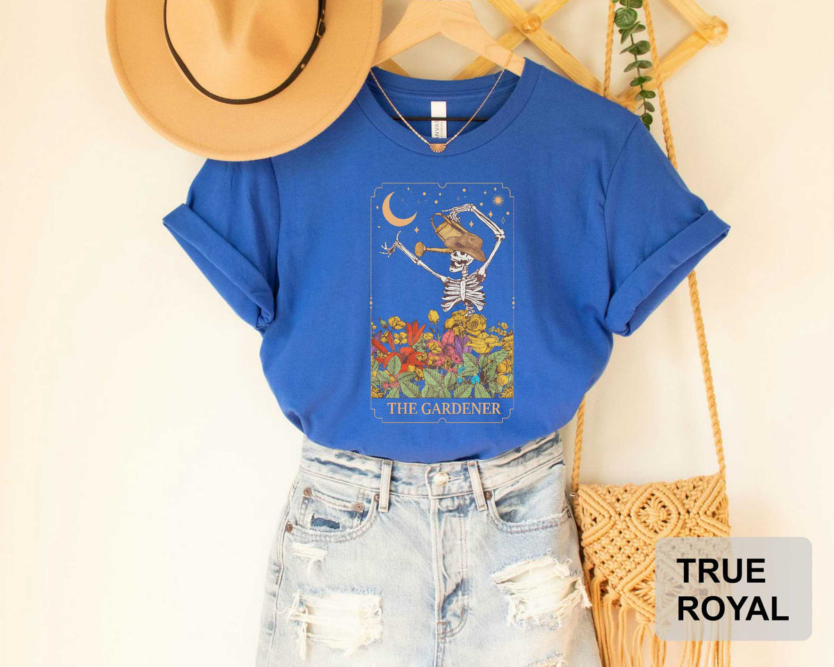 A blue t-shirt with a graphic design of a skeleton holding a flower, along with a beige hat and a woven purse, all displayed on a hanger.