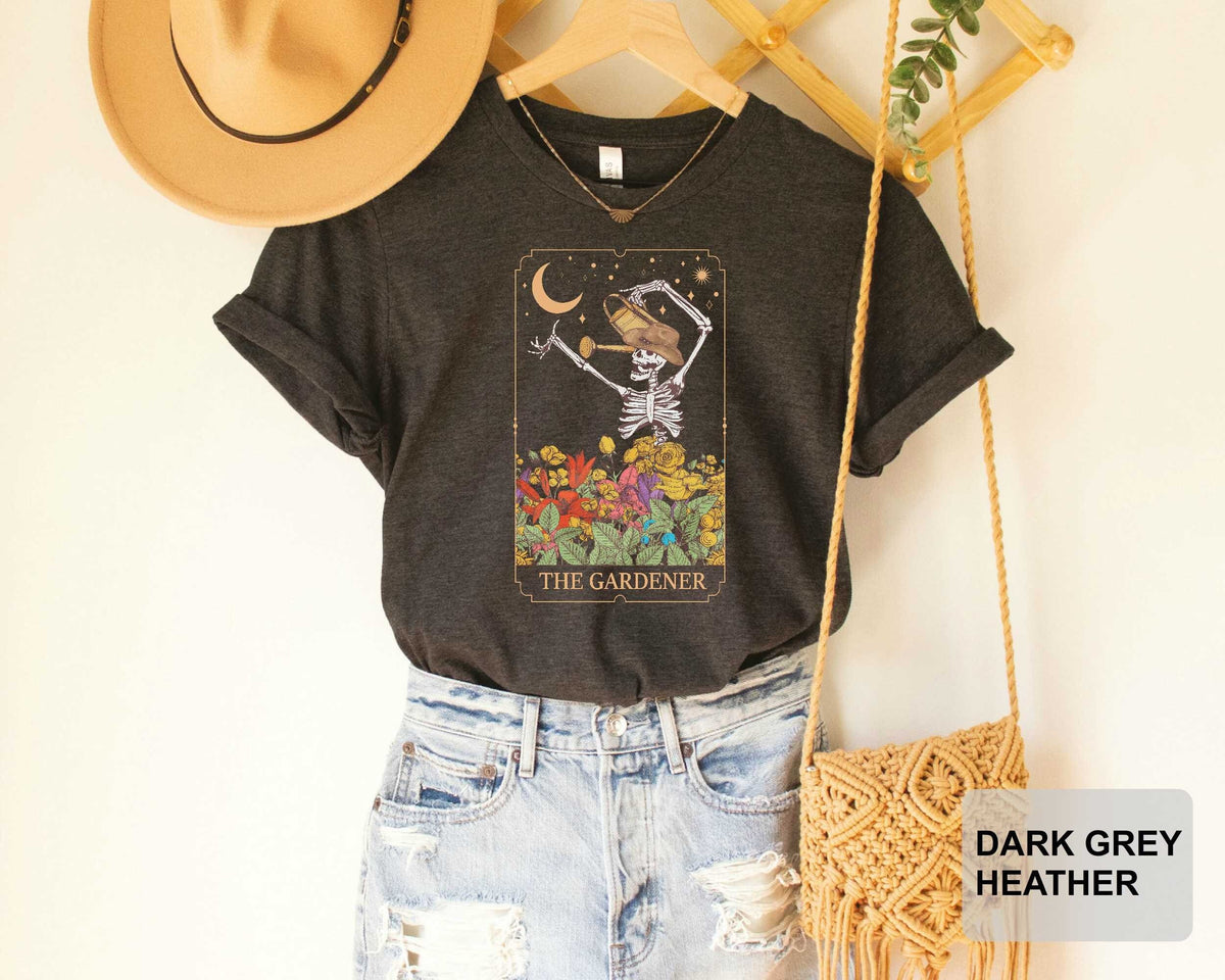 A black t-shirt with a graphic design of a skeleton holding a flower, along with the text "THE GARDENER", is displayed on a hanger. The t-shirt is paired with distressed denim jeans and a beige hat. A woven purse is also visible in the image.