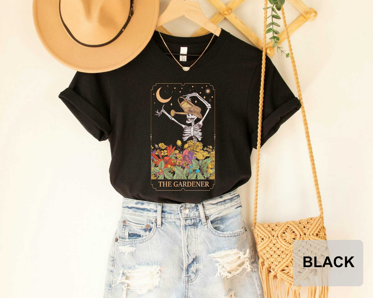 a black t-shirt with a graphic of a skeleton holding a watering can, along with the text "THE GARDENER" printed on it. The t-shirt is paired with distressed denim jeans and a straw hat, and a woven purse is also visible in the scene.