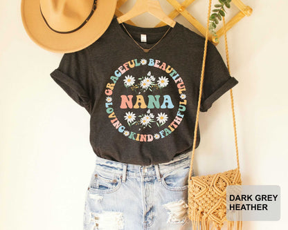 A black t-shirt with a colorful floral design and the word "NANA" printed on it, along with the text "NOT GRACEFUL, BEAUTIFUL, BEAUTIFUL, KIND, FAITHFUL" arranged around it. The t-shirt is hanging on a wooden hanger, and there is a beige hat and a woven purse nearby.
