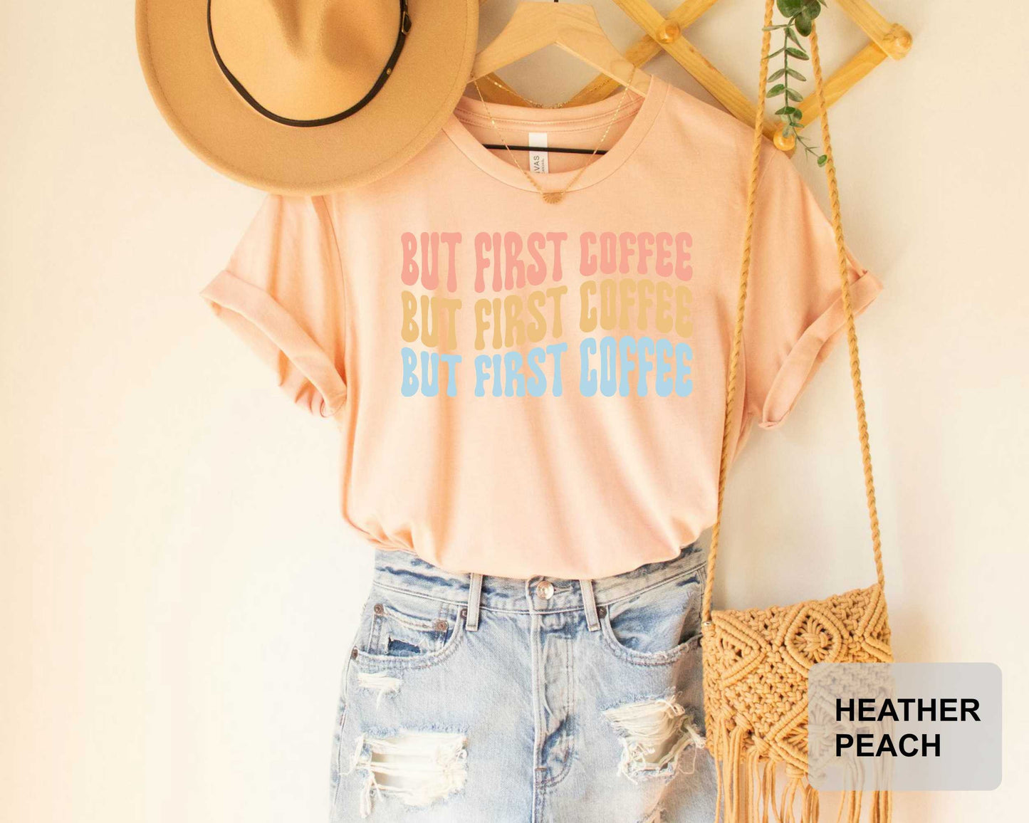 A casual outfit consisting of a peach-colored t-shirt with the text "BUT FIRST COFFEE BUT FIRST COFFEE BUT FIRST COFFEE" printed on it, paired with distressed denim jeans and a straw hat. The outfit is displayed on a wooden hanger, with a woven purse hanging nearby.