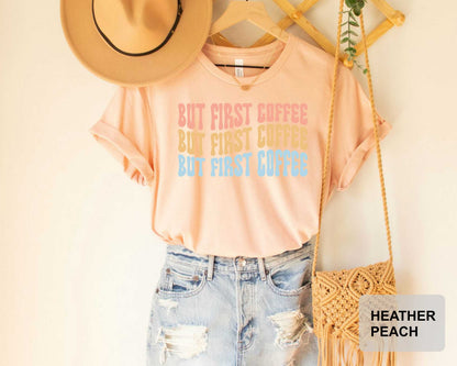 A casual outfit consisting of a peach-colored t-shirt with the text "BUT FIRST COFFEE BUT FIRST COFFEE BUT FIRST COFFEE" printed on it, paired with distressed denim jeans and a straw hat. The outfit is displayed on a wooden hanger, with a woven purse hanging nearby.