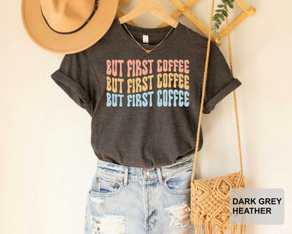 A gray t-shirt with the text "BUT FIRST COFFEE BUT FIRST COFFEE BUT FIRST COFFEE" printed on it, along with a pair of distressed denim jeans and a beige hat. The t-shirt is hanging on a wooden hanger, and there is a woven purse nearby.