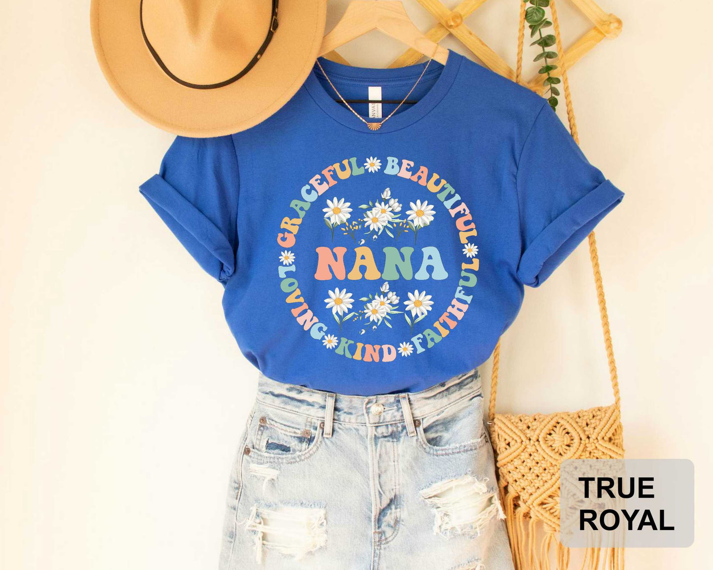 A blue t-shirt with a floral design and the word "NANA" printed on it, along with a pair of denim shorts and a beige hat, are displayed on a hanger.